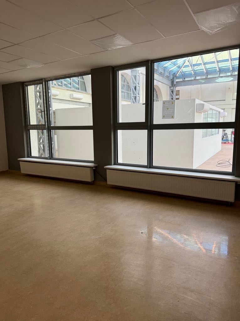 Puste, jasne pomieszczenie z dwoma dużymi oknami wychodzącymi na dziedziniec z przeszklonym dachem, widoczne grzejniki pod oknami, podłoga w odcieniach beżu.