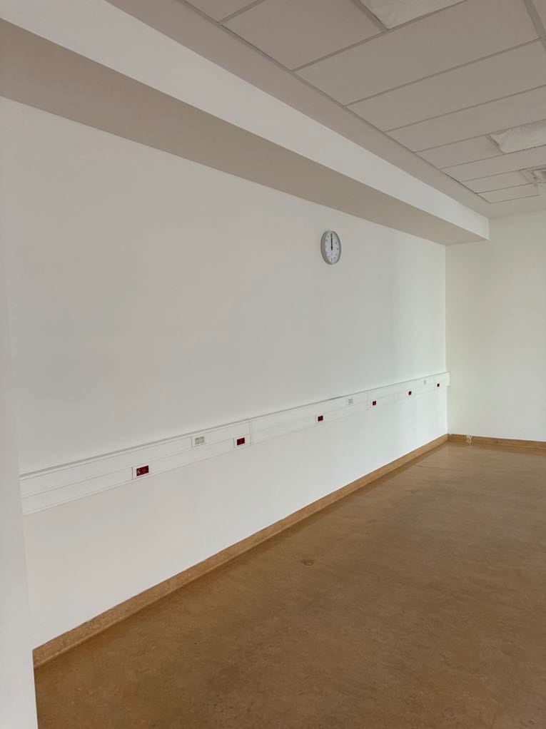 Wnętrze wykończonego pomieszczenia z białymi ścianami, brązową podłogą, listwą przypodłogową i zegarem na ścianie. Widoczne gniazdka elektryczne w listwie. Sufit podwieszany.