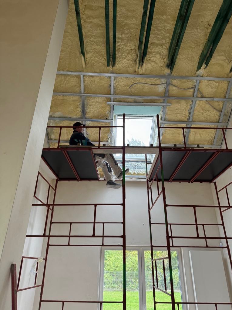 Pracownik na rusztowaniu, widoczne ocieplenie poddasza pianą, konstrukcja metalowa pod płyty, okno dachowe