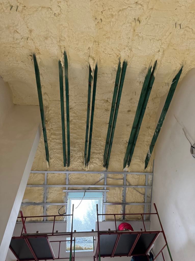 Montaż płyty karton gips na poddaszu z izolacją pianką PUR, widoczne zielone belki konstrukcyjne i rusztowanie.