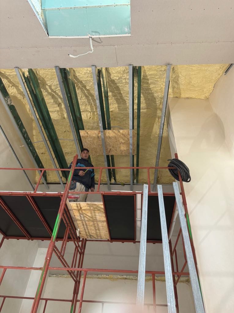 Pracownik na rusztowaniu montuje konstrukcję sufitu podwieszanego z izolacją piankową, widoczne profile metalowe i płyta.