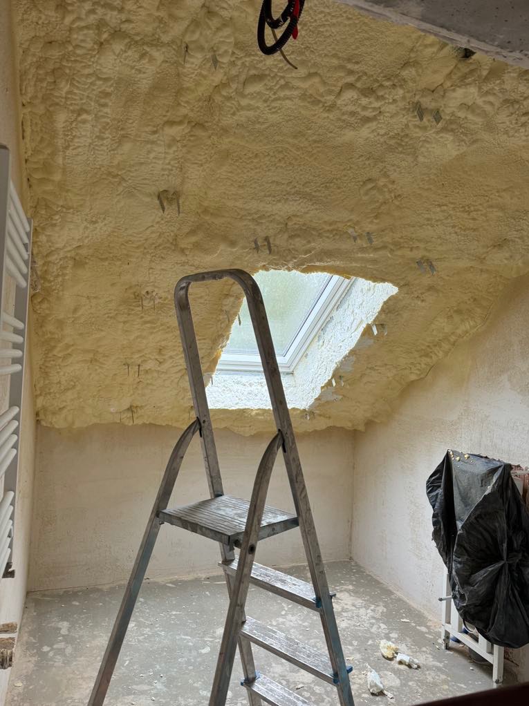 Poddasze ocieplone pianą poliuretanową, widoczna drabina aluminiowa i okno dachowe, fragment instalacji elektrycznej pod sufitem, folia ochronna na ścianie.