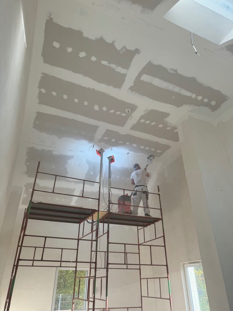 Pracownik na rusztowaniu szlifuje sufit z zamontowanymi płytami, przygotowany do dalszych prac wykończeniowych. Widoczne lampy halogenowe oświetlające miejsce pracy i odkurzacz przemysłowy. Sufit...