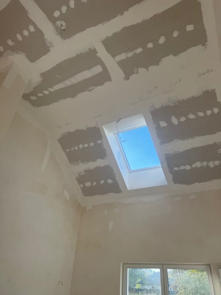 Wnętrze w trakcie prac wykończeniowych: sufit z widocznymi łączeniami płyt kartonowo-gipsowych i wstępnie położoną warstwą szpachli, okno dachowe z widokiem na błękitne niebo, ściany przygotowane...
