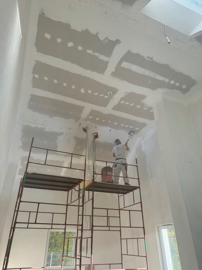 Pracownik szlifuje połączenia płyt na suficie podwieszanym z płyty gipsowo-kartonowej, stojąc na rusztowaniu w wysokim pomieszczeniu z oknem.