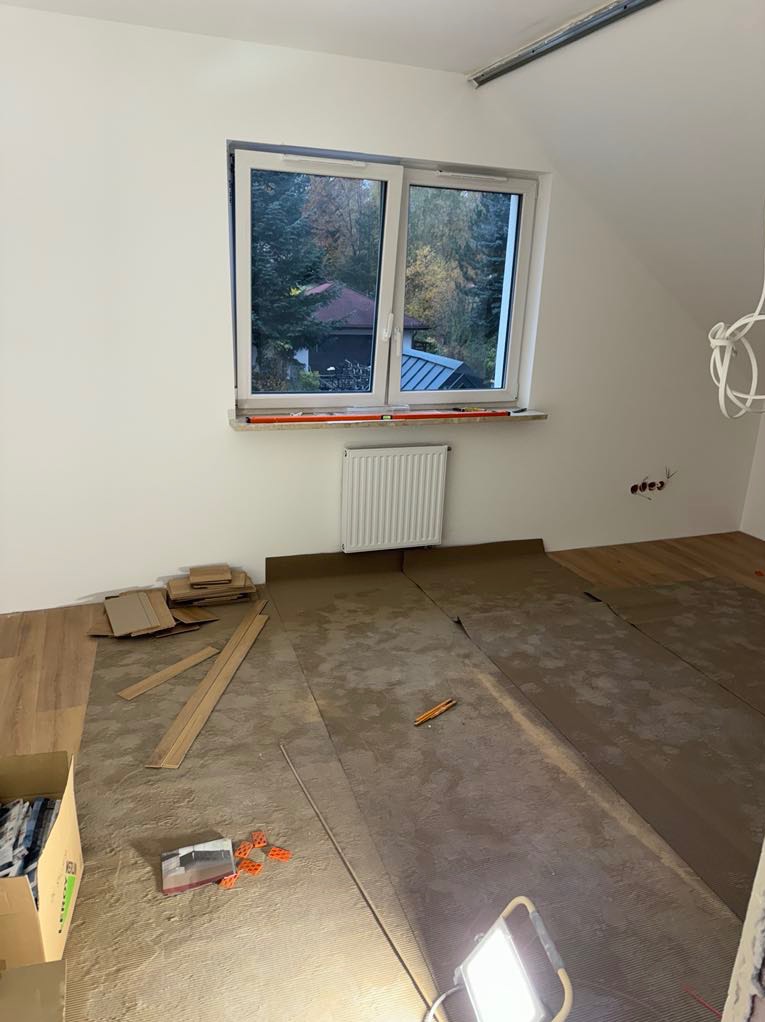 Pokój na poddaszu w trakcie montażu paneli podłogowych; widoczne rozłożone panele, folia izolacyjna, kartony, poziomica na parapecie oraz lampa budowlana oświetlająca podłogę.
