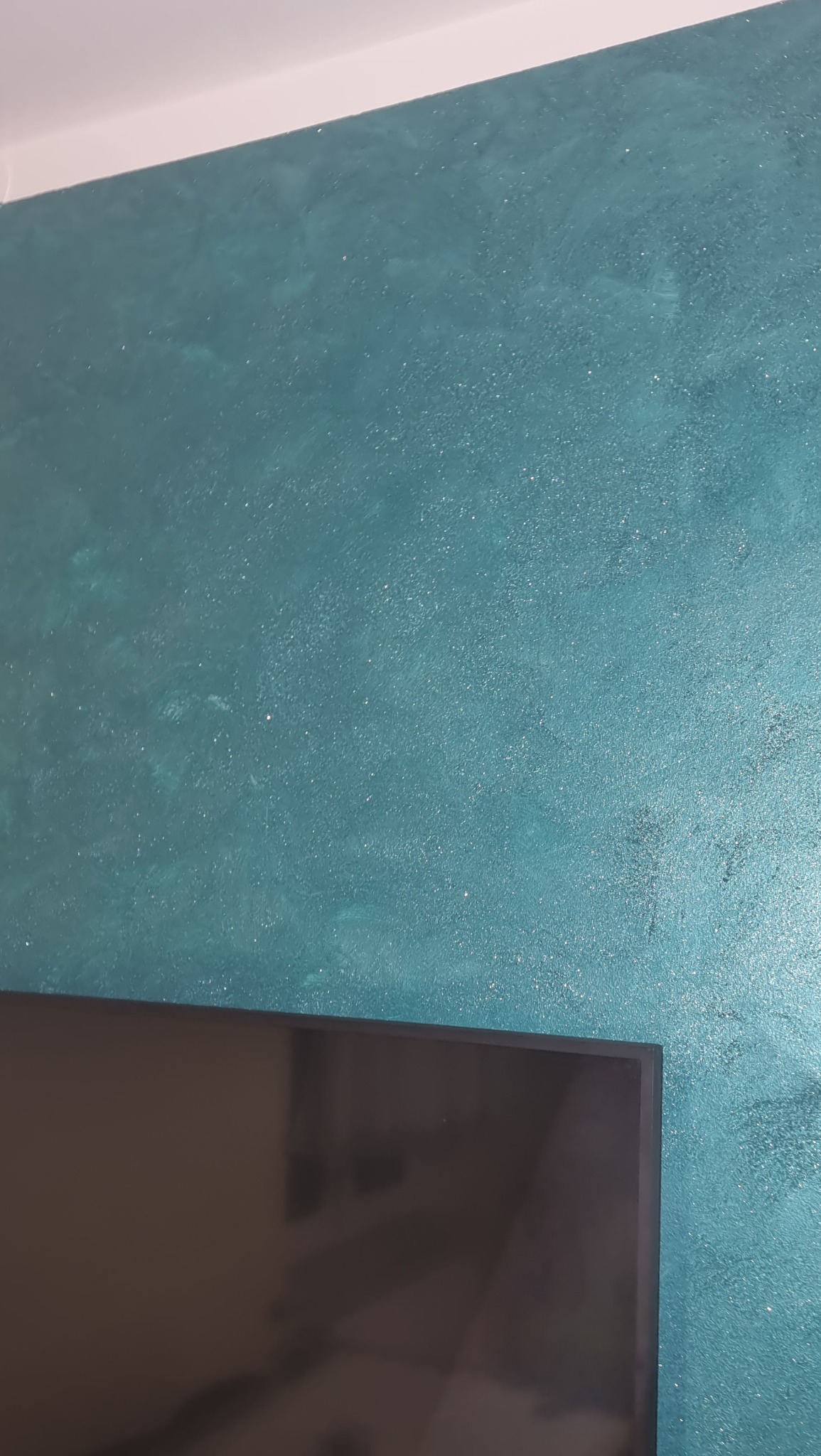 Turkusowa ściana z dekoracyjną, połyskującą fakturą, fragment telewizora w dolnej części kadru.