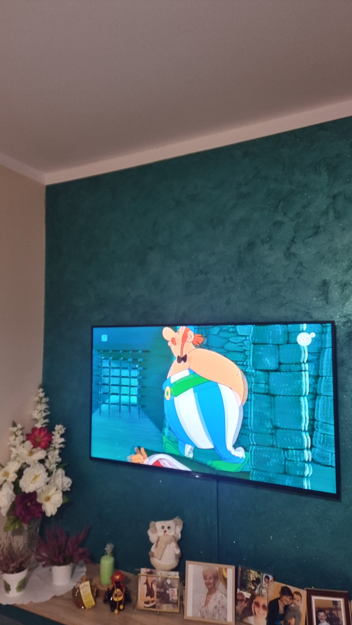 Ściana w kolorze butelkowej zieleni z dekoracyjną fakturą, podświetlona telewizorem z włączoną kreskówką Asterix i Obelix, na szafce pod telewizorem kwiaty w wazonie i ramki ze zdjęciami.