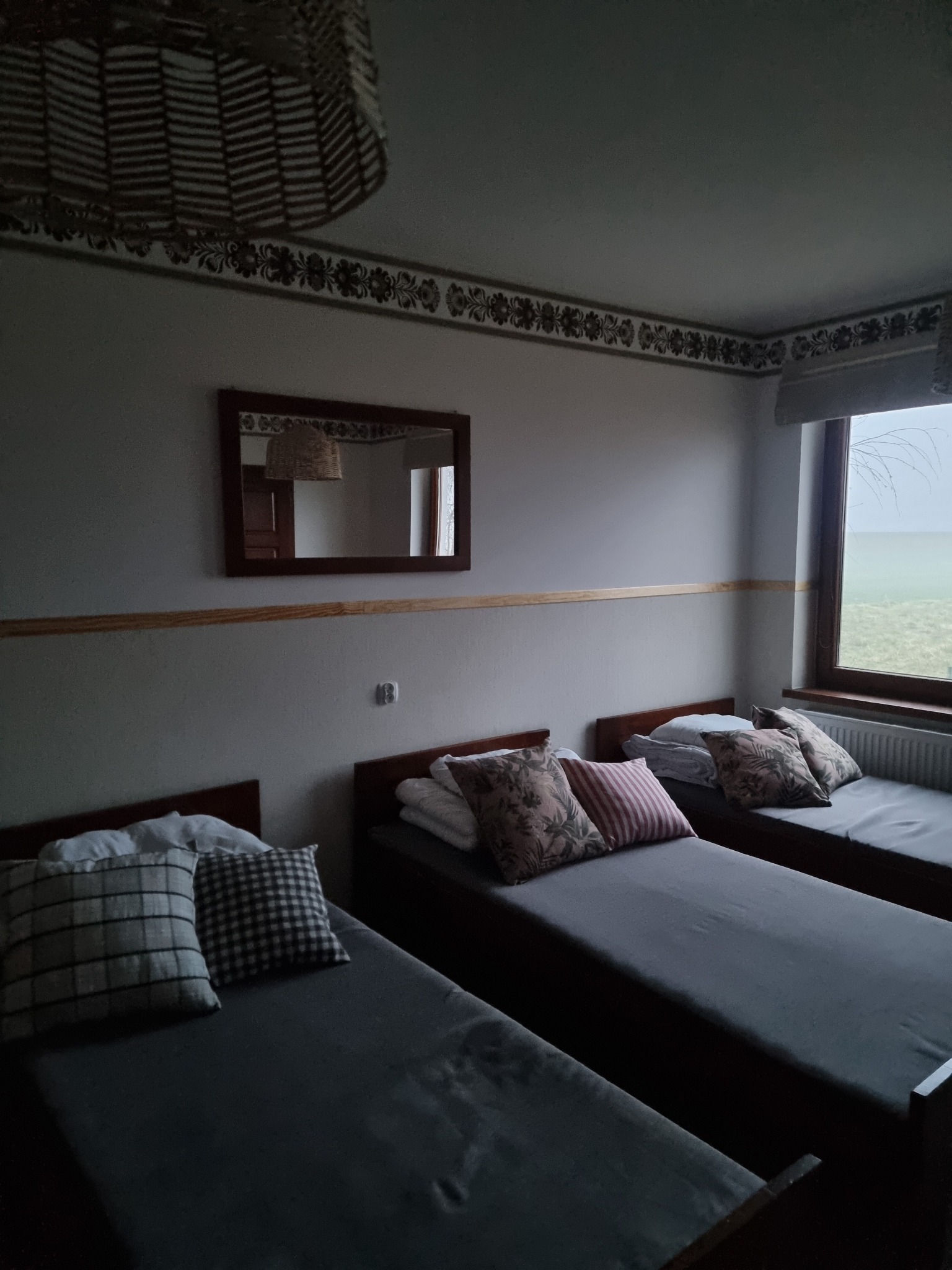 Pokój z trzema łóżkami jednoosobowymi, szarymi narzutami i różnymi poduszkami, widok z okna na zewnątrz, dekoracyjna listwa pod sufitem i lampa z wikliny.