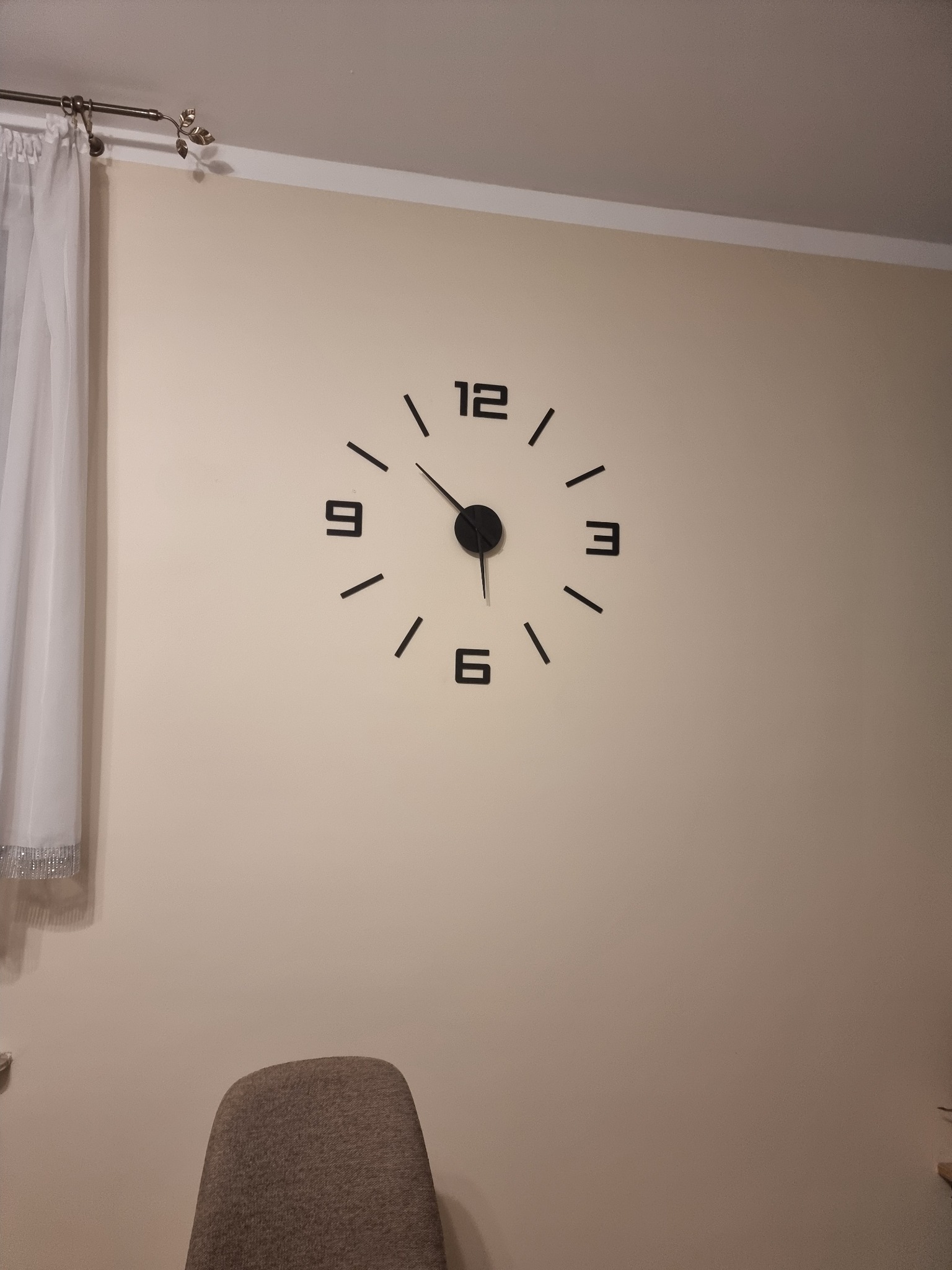 Minimalistyczny zegar ścienny z czarnymi cyframi i wskazówkami na beżowej ścianie, widoczny fragment zasłony i krzesła, oświetlenie górne.