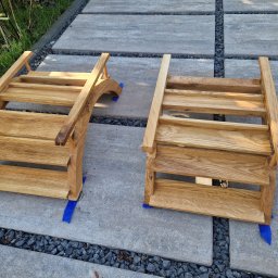 Tymoteusz Berny - Dwa drewniane podesty schodkowe, wykonane z jasnego drewna, ustawione na betonowych płytach z kamienną podsypką; jeden z podestów ma zaokrąglony bok, oba zabezpieczone niebieską taśmą.