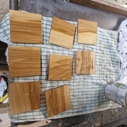 Tymoteusz Berny - Kwadratowe elementy z drewna o widocznym usłojeniu, częściowo polakierowane, leżące na niebiesko-białej ścierce w warsztacie stolarskim, obok pędzla i puszki z lakierem.