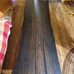 Tymoteusz Berny - Długi, ciemny blat z litego drewna o nieregularnych krawędziach, położony na drewnianej podłodze. Widoczne słoje drewna i naturalne pęknięcia. W tle fragment komody i dywanu.