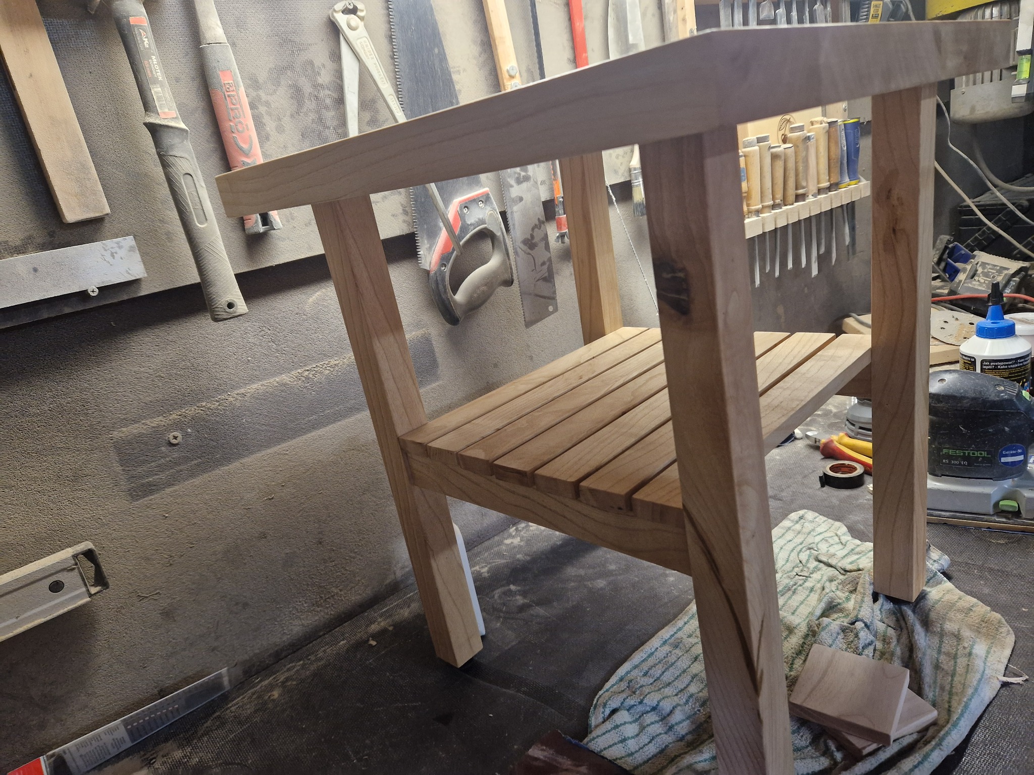 Niedokończony, drewniany stolik nocny z półką, widoczny w warsztacie stolarskim, z narzędziami na ścianie i frezarką Festool w tle.
