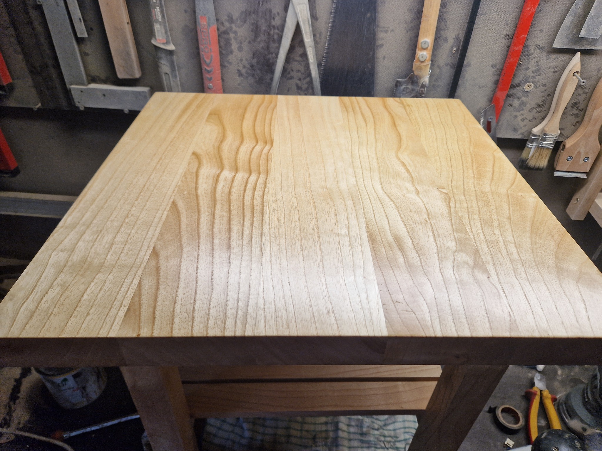 Kwadratowy blat z jasnego drewna, na tle warsztatu z narzędziami, widoczne słoje drewna i fragment konstrukcji nośnej.