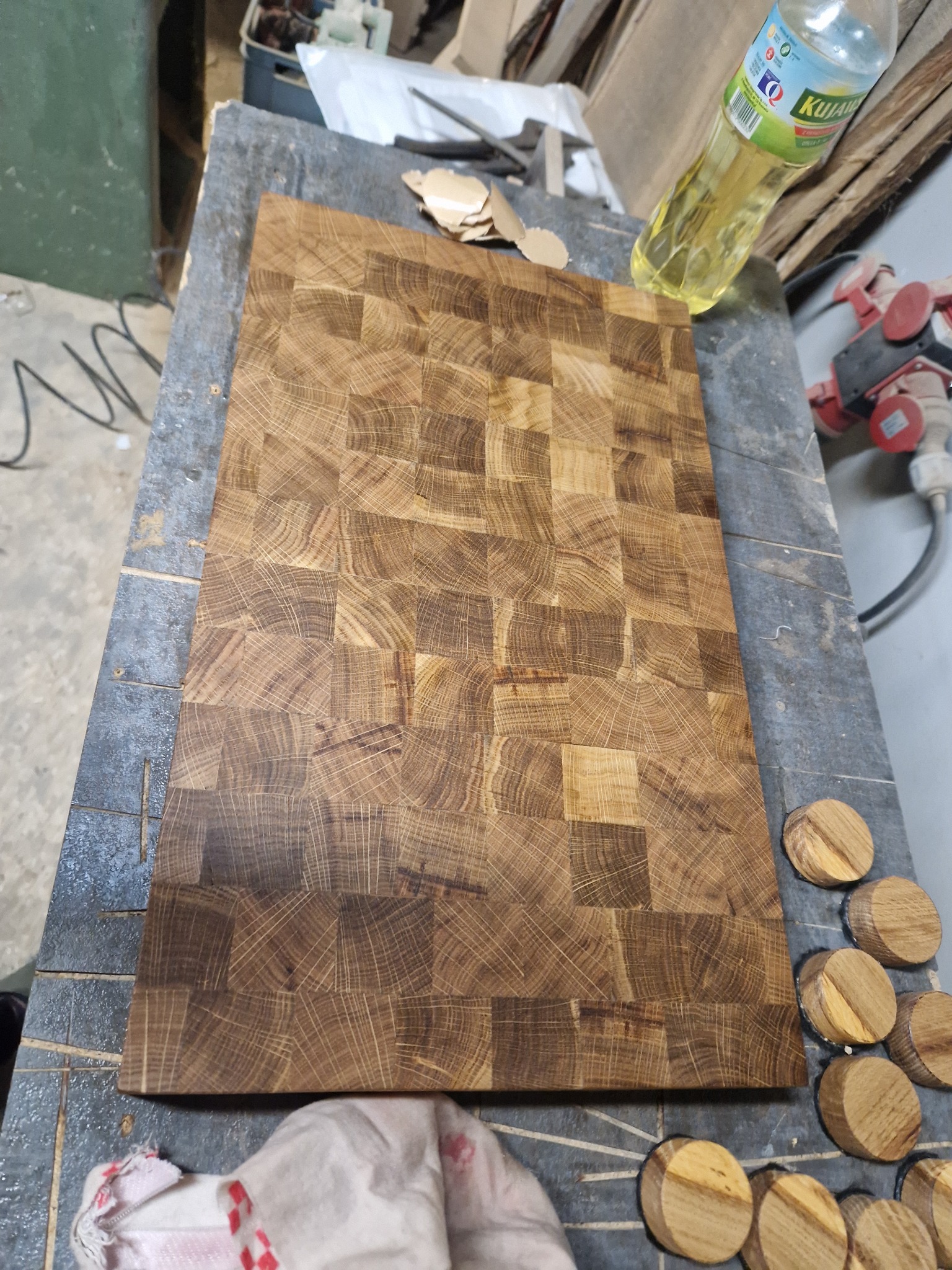 Surowy blat drewniany z mozaiką z kwadratowych klocków, leżący na warsztatowym stole, obok widoczne niedokończone elementy i narzędzia, sugerujące etap produkcji.