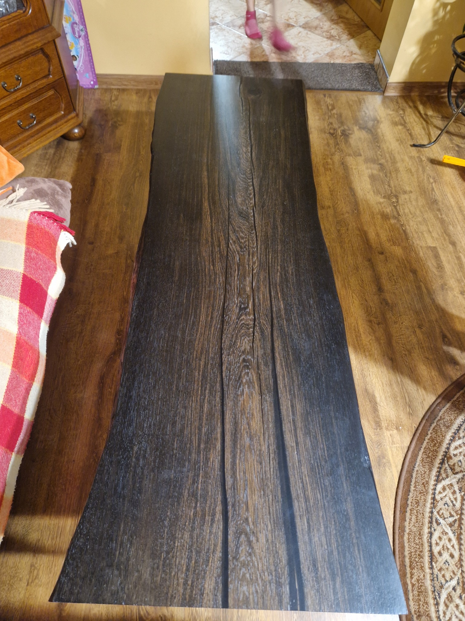 Długi, ciemny blat z litego drewna o nieregularnych krawędziach, położony na drewnianej podłodze. Widoczne słoje drewna i naturalne pęknięcia. W tle fragment komody i dywanu.