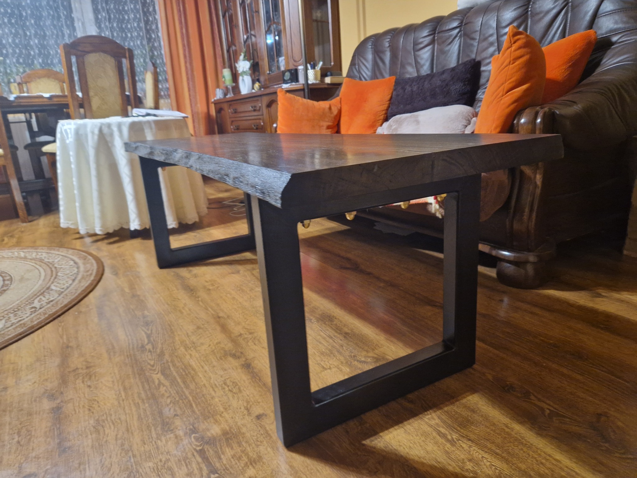 Niski stolik kawowy z ciemnego drewna z metalowymi nogami w kształcie kwadratów, na tle brązowej skórzanej kanapy z pomarańczowymi poduszkami i drewnianych mebli.