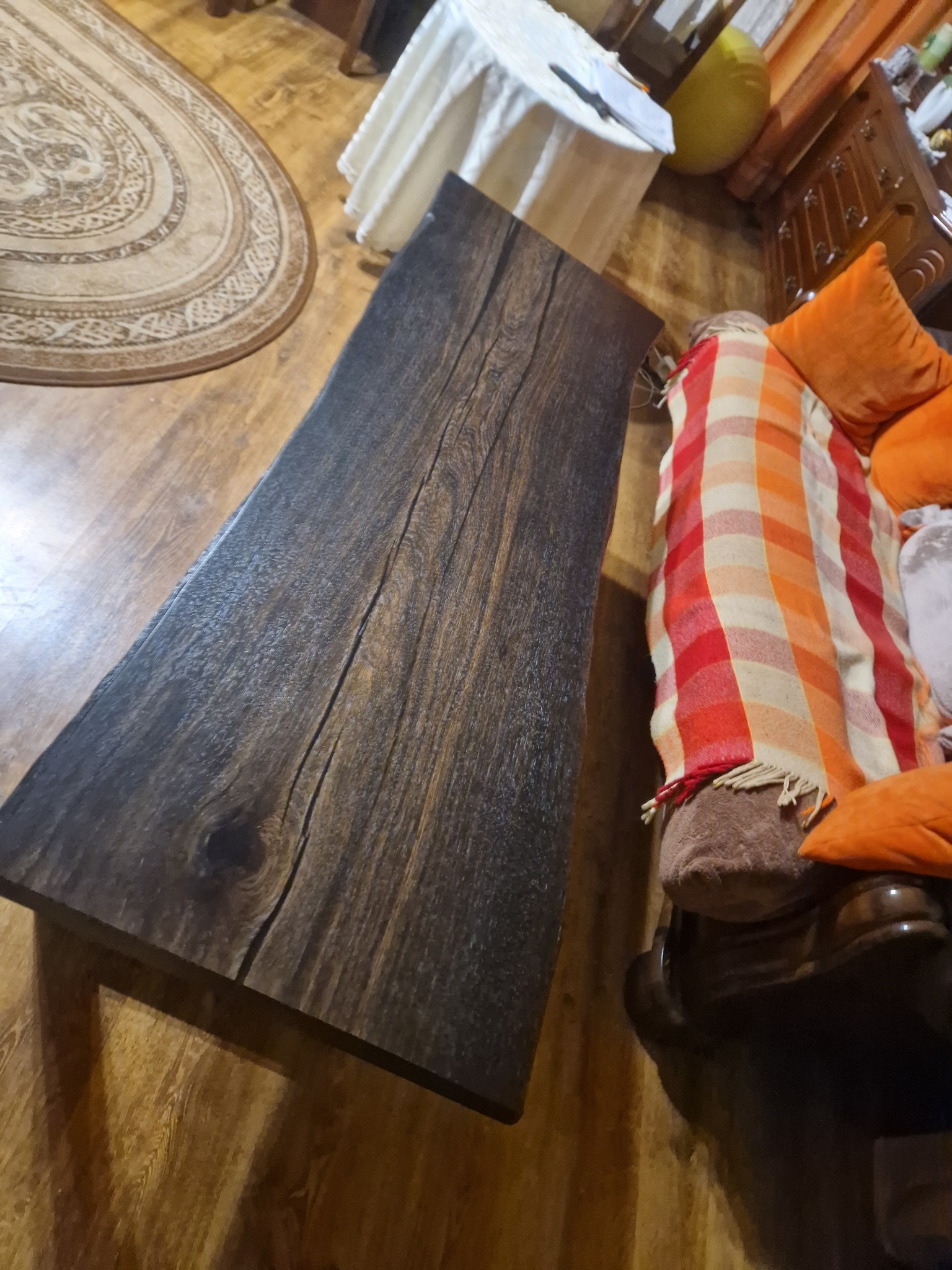 Nieregularny stolik kawowy z ciemnego drewna z wyraźnym usłojeniem, ustawiony w salonie obok sofy z pomarańczowymi poduszkami i pledem w kratę.