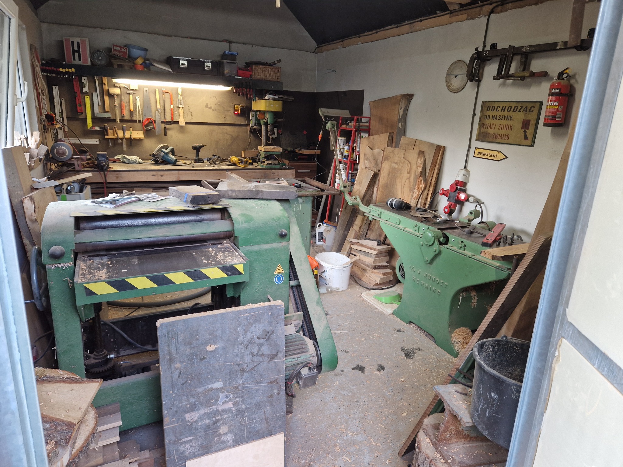 Warsztat stolarski z widocznymi strugarkami, narzędziami na ścianie i stosem drewna. Na ścianie tablica informacyjna i gaśnica.