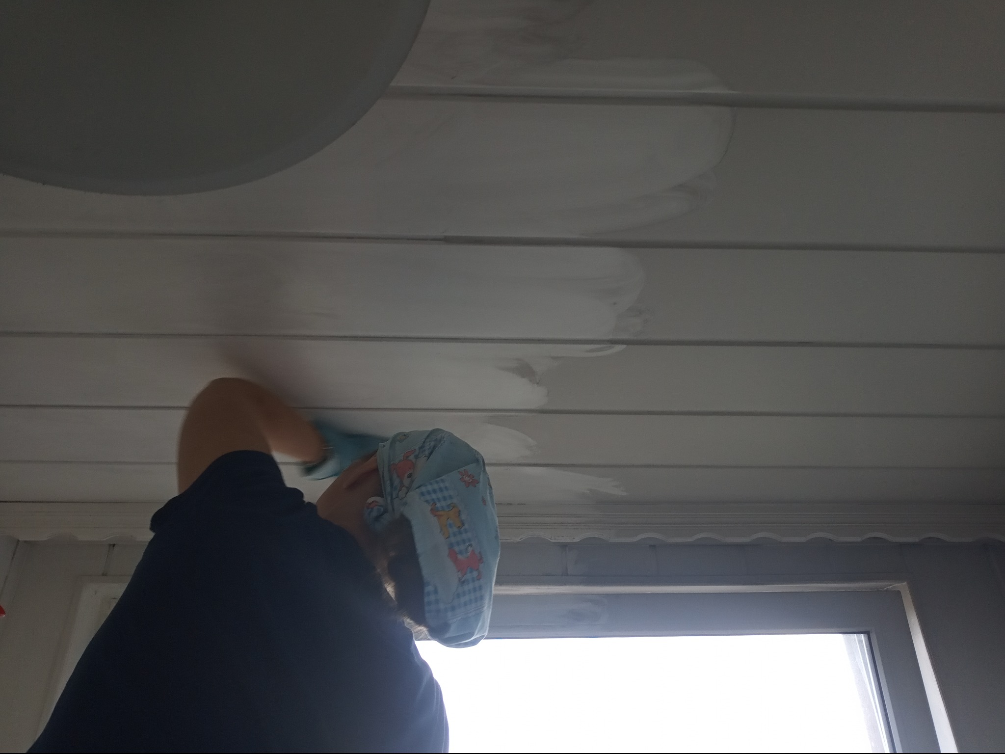 Osoba w czepku ochronnym czyści biały sufit z paneli, widoczne ślady po czyszczeniu, okno w tle.