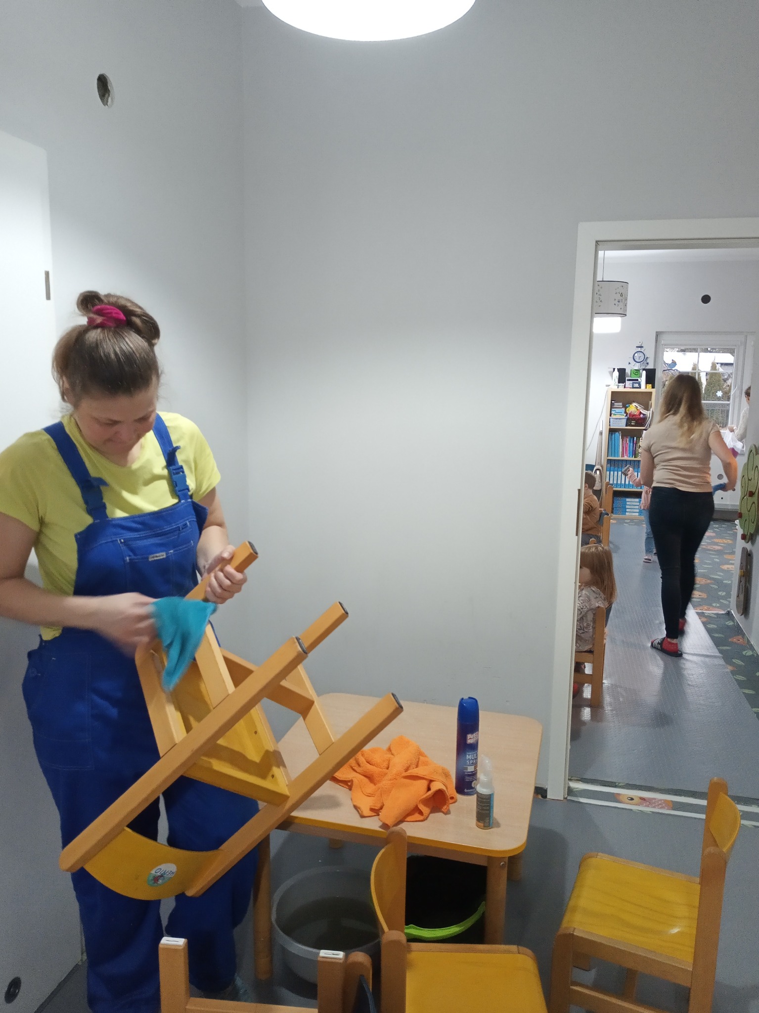 Kobieta w niebieskim kombinezonie czyści wilgotną szmatką krzesło w przedszkolu, na stole środki czystości, w tle widoczne dzieci i druga kobieta.