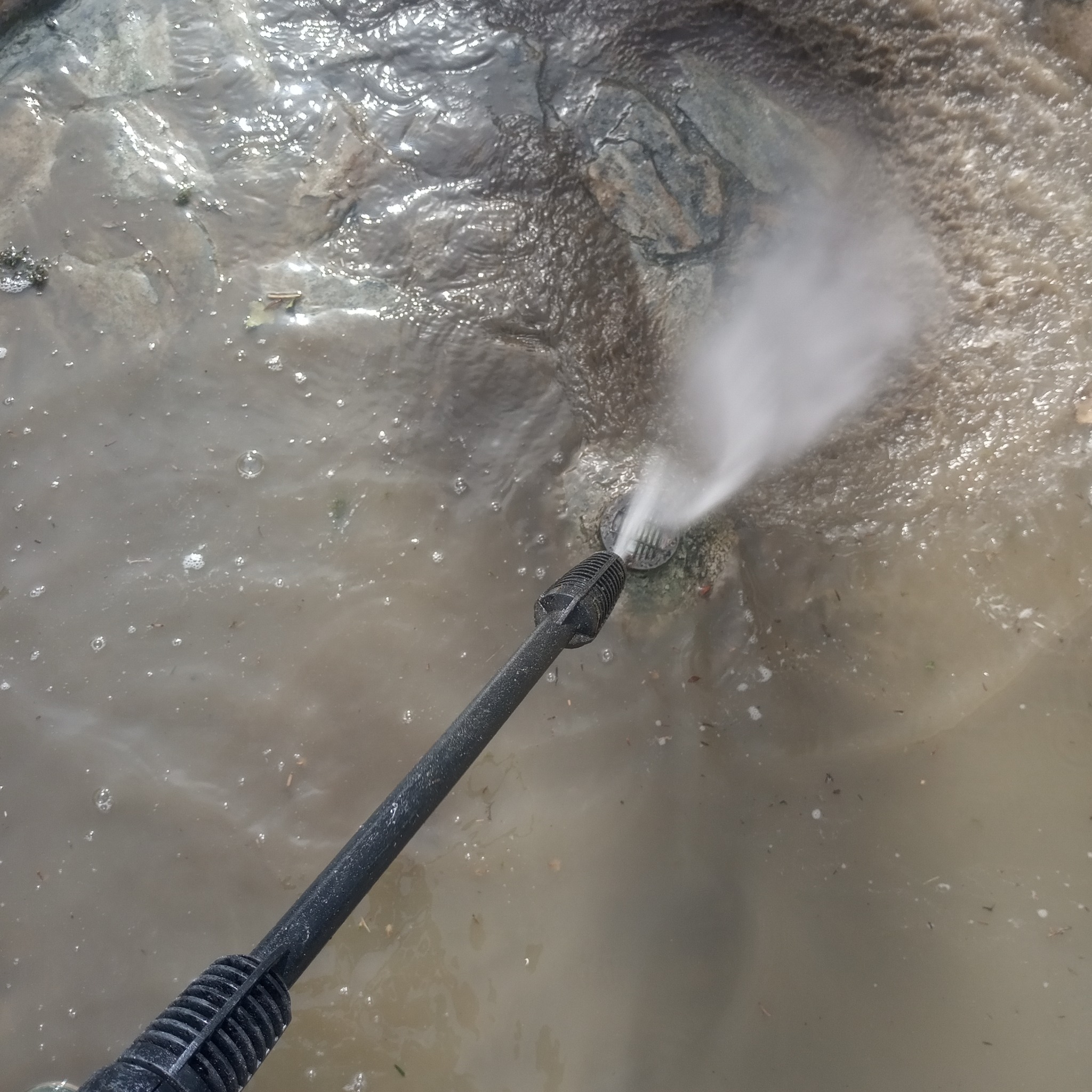 Czyszczenie odpływu kanalizacyjnego z osadów za pomocą myjki ciśnieniowej, widoczny strumień wody i zabrudzona woda wokół.