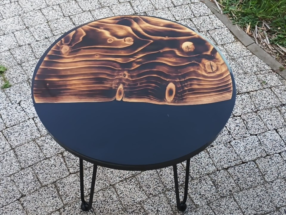 Okrągły stolik kawowy z blatem z opalizowanego drewna i granatową podstawą, stojący na trzech metalowych nogach typu hairpin, na tle kostki brukowej.