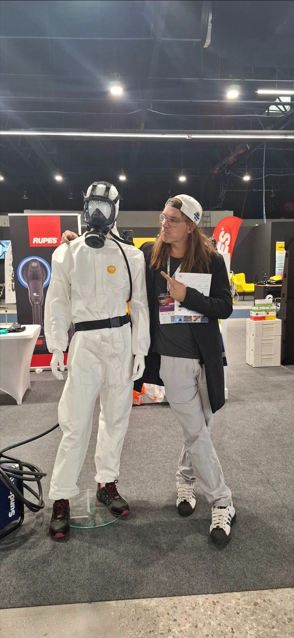 Mężczyzna w czapce wskazuje na manekina w kombinezonie ochronnym i masce gazowej na tle stoiska z polerkami Rupes na targach.