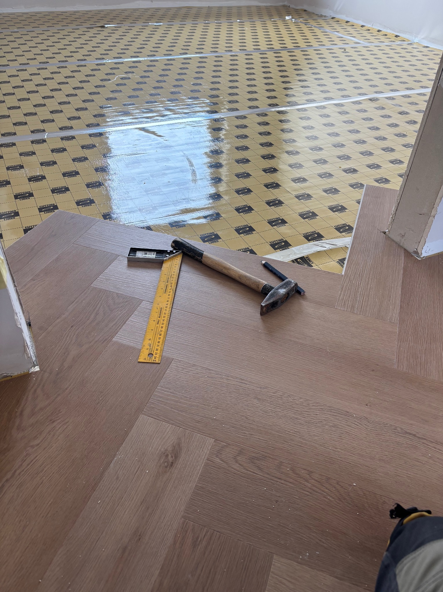 Fragment nowej podłogi ułożonej w jodełkę, widoczne narzędzia: młotek, kątownik i ołówek, pod spodem folia izolacyjna z nadrukiem.
