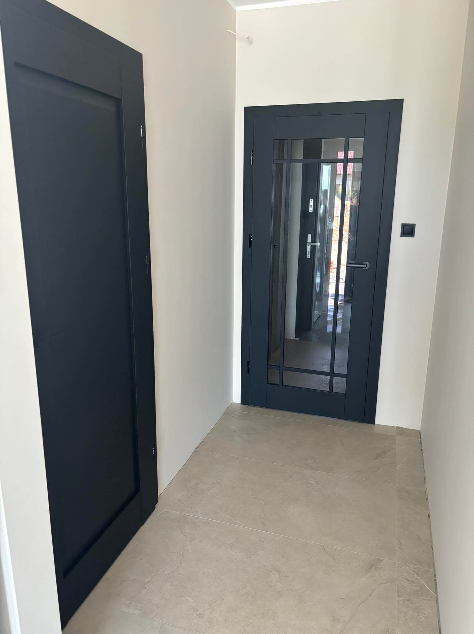 Dwa ciemnoszare skrzydła drzwiowe w jasnym korytarzu, jedno pełne, drugie z szybami w czarnej ramie, widok na wnętrze domu przez szklane panele.