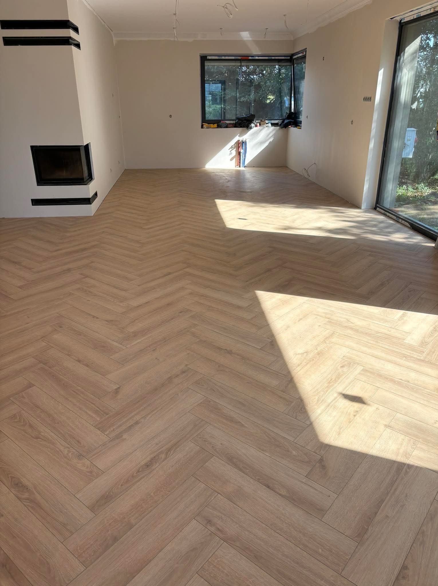 Jasne, drewnopodobne panele ułożone w jodełkę w przestronnym, jasnym pokoju z minimalistycznym kominkiem i dużym oknem.