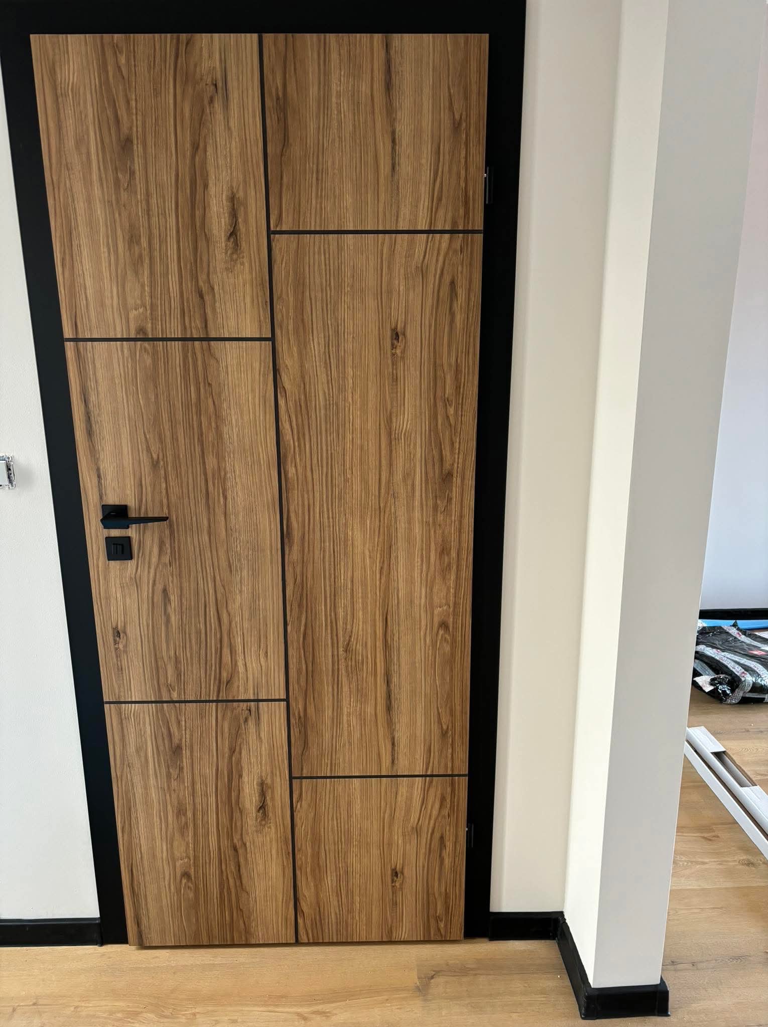 Nowoczesne drzwi wewnętrzne z okleiną drewnopodobną w odcieniach brązu, z czarną ościeżnicą i minimalistyczną klamką, zamontowane w jasnym pomieszczeniu z drewnianą podłogą.