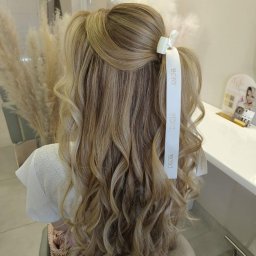 sTyLove Upięcia Studio Pielęgnacji Włosów EWELINA STANKIEWICZ - Upięcie blond włosów z lokami, ozdobione białą wstążką Dior, widok z tyłu na tle dekoracji z traw pampasowych.