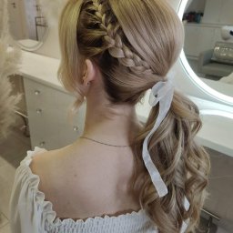 sTyLove Upięcia Studio Pielęgnacji Włosów EWELINA STANKIEWICZ - Blond włosy upięte w wysoki kucyk z warkoczem i białą wstążką, widok z tyłu na tle jasnego wnętrza salonu.