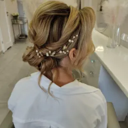 Upięcie ślubne z blond włosów z ozdobną spinką z pereł i kamieni, widok z tyłu, biała koszula, jasne wnętrze salonu.