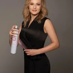 Kobieta w czarnej sukience spryskuje włosy lakierem, trzymając w drugiej ręce różowy grzebień na szarym tle.