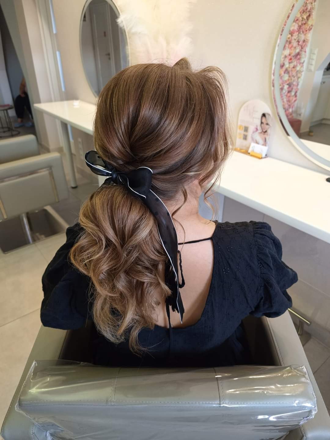 Upięcie z brązowych włosów z lokami, upięte czarną wstążką w kok, widok z tyłu na tle salonu fryzjerskiego.
