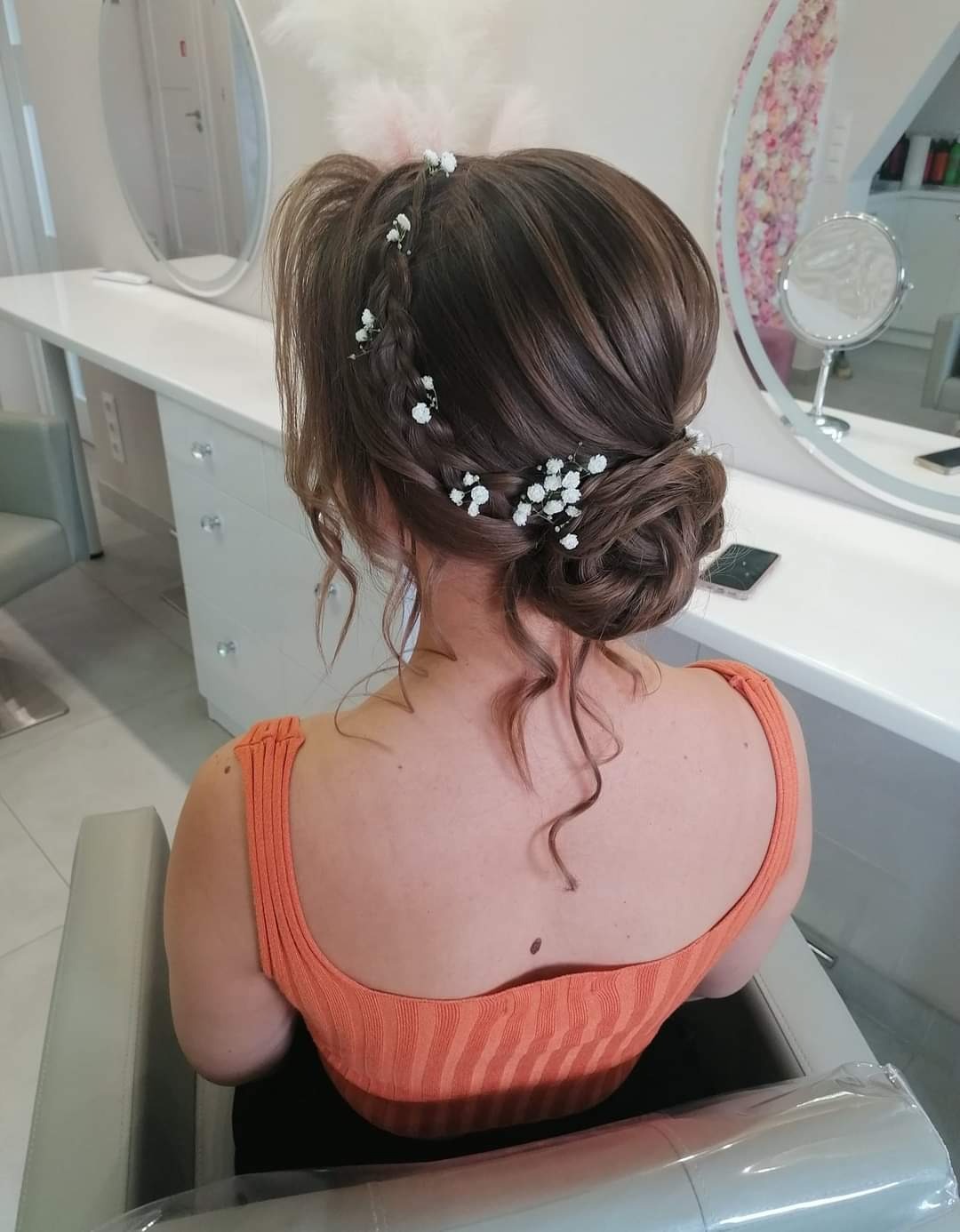 Upięcie włosów z warkoczem i drobnymi białymi kwiatami, widok z tyłu, w tle lustro i białe meble salonu.