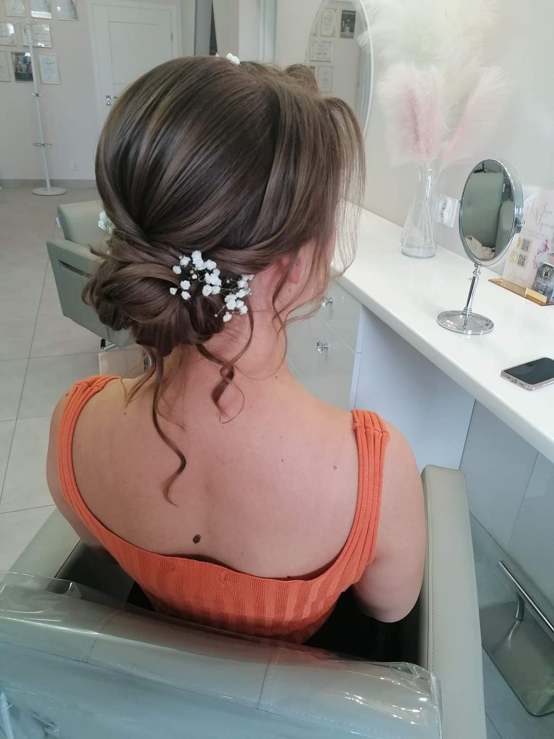Upięcie włosów z wplecionymi drobnymi, białymi kwiatkami, widok z tyłu na upiętą fryzurę kobiety w pomarańczowej bluzce, siedzącej w fotelu w salonie.