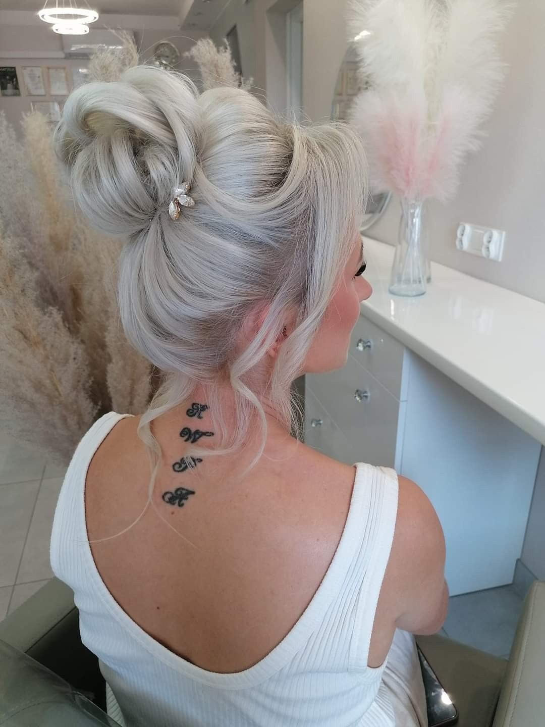 Elegancka fryzura upięta w kok z ozdobną spinką, włosy w odcieniach platynowego blondu, widoczny tatuaż na karku kobiety.