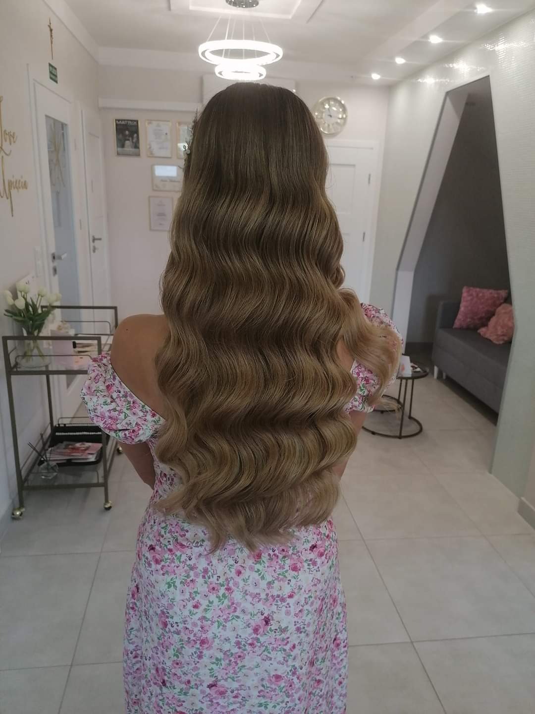Długie, falowane włosy w odcieniach blondu i brązu, widziane z tyłu, na kobiecie w sukience w kwiaty, w jasnym wnętrzu salonu.