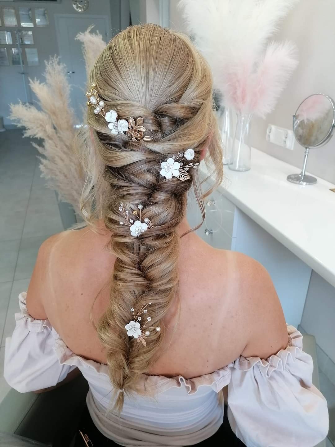 Blond włosy upięte w gruby warkocz ozdobiony białymi kwiatami i złotymi listkami, widok z tyłu, odsłonięte ramiona w bluzce z falbankami.