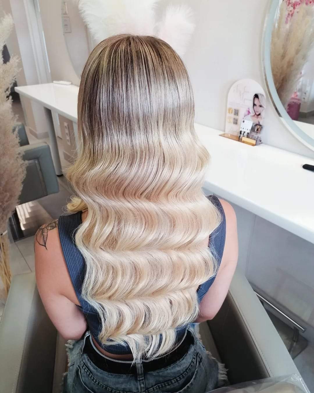 Blond włosy z delikatnymi falami od tyłu, z widocznym ombre od ciemnego blondu u nasady po jasny blond na końcach, w salonie z białymi meblami.