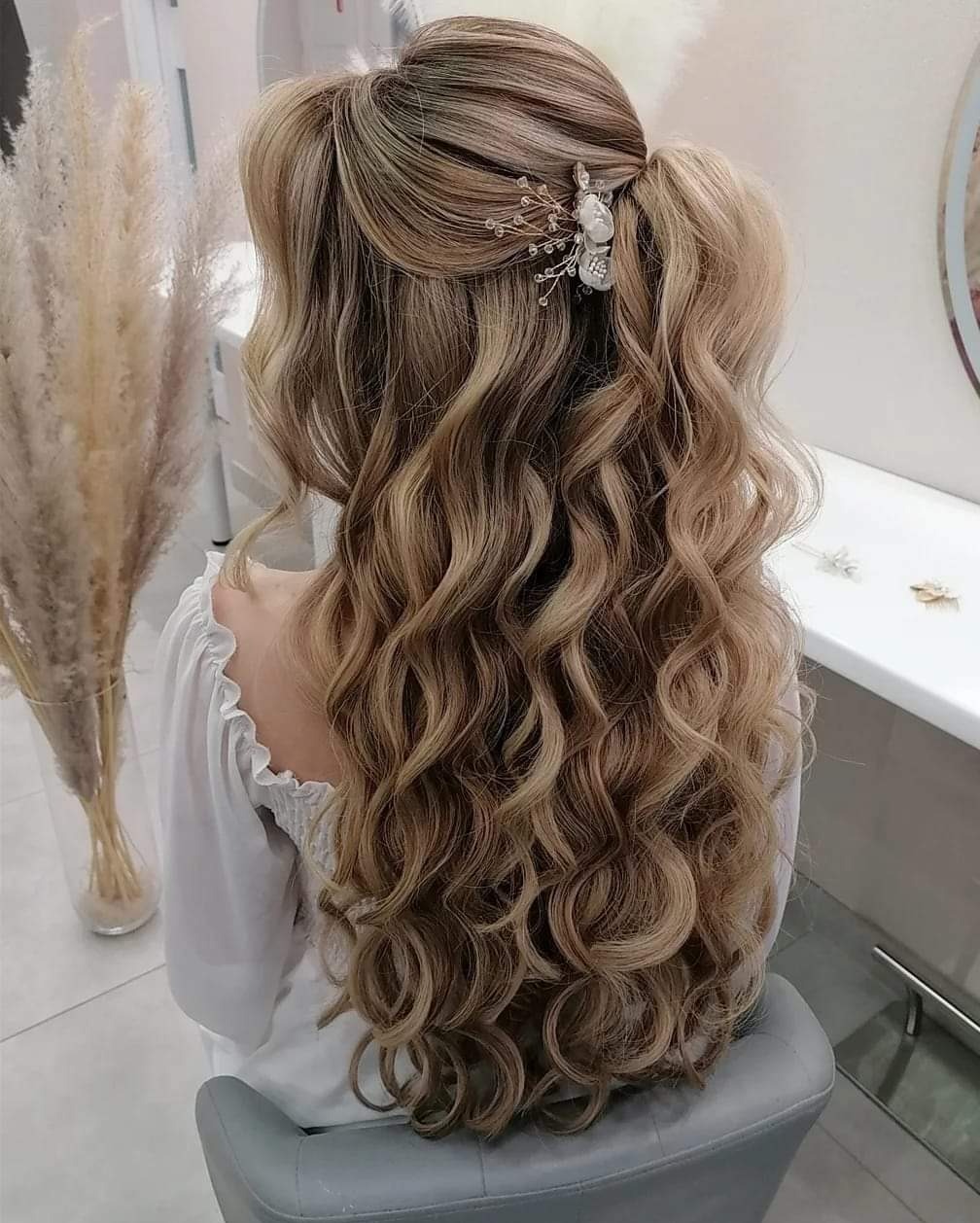 Upięcie z loków blond i brąz, spięte ozdobną spinką z kwiatami, widok z tyłu na tle jasnego wnętrza salonu.