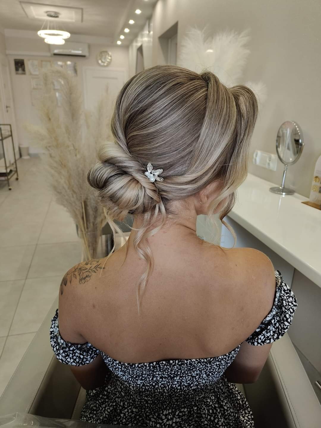 Elegancka upięta fryzura blond z wplecionym ozdobnym motywem kwiatowym, widok z tyłu, odsłonięte ramiona, tatuaż na ramieniu, salon fryzjerski w tle.