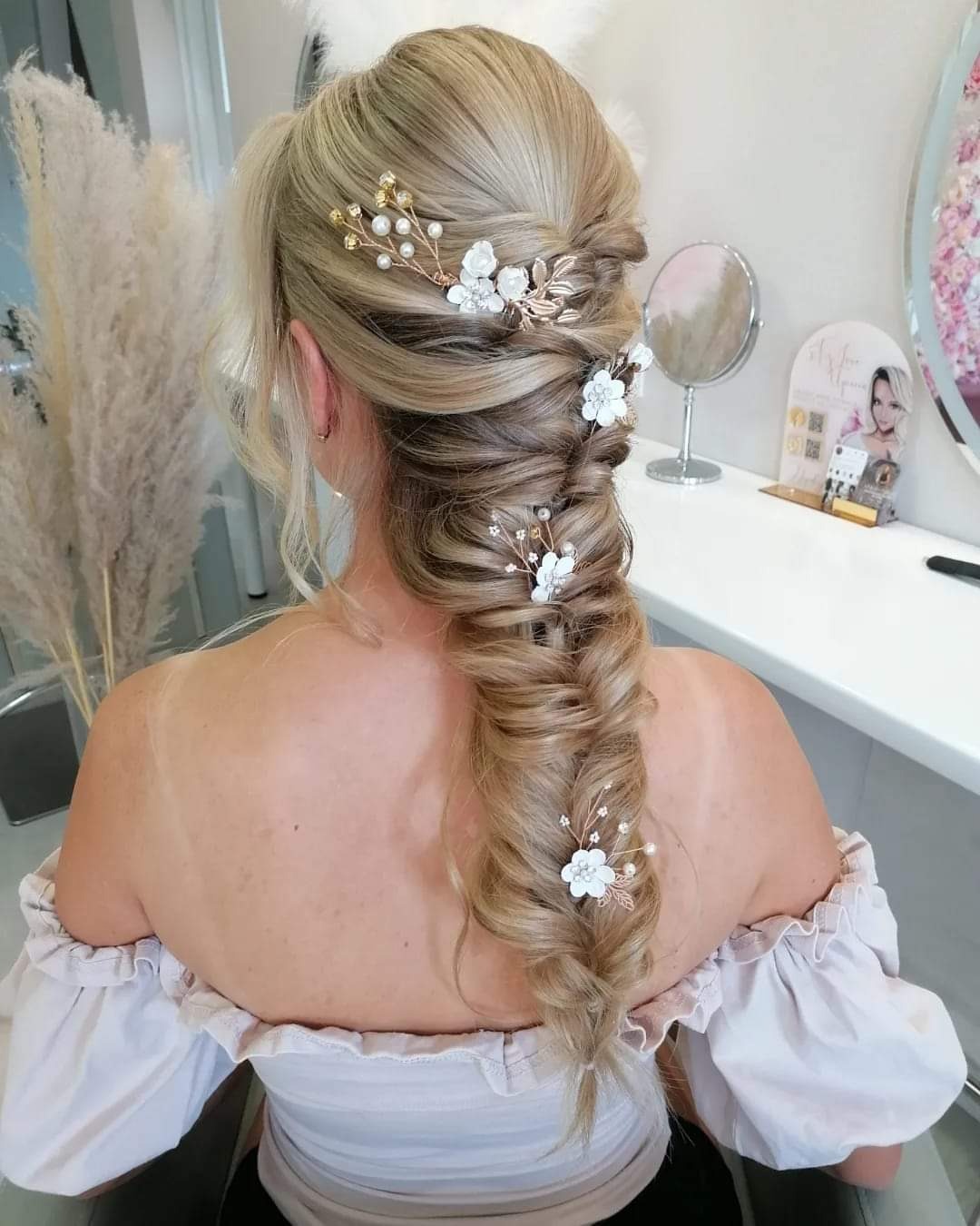 Blond włosy upięte w fantazyjny warkocz typu rybi ogon, ozdobione białymi kwiatami i złotymi akcentami, widok z tyłu na tle salonu.
