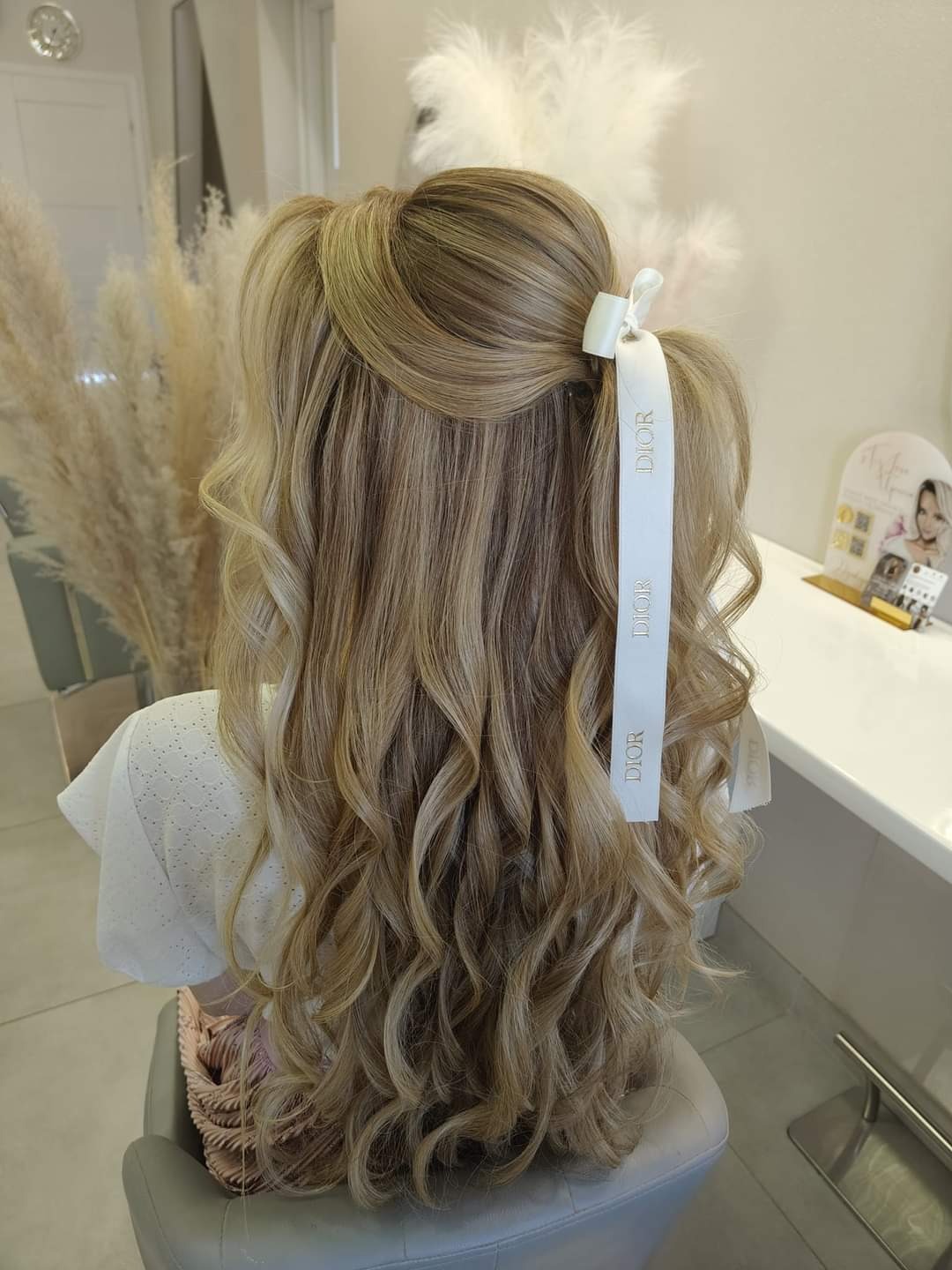 Upięcie blond włosów z lokami, ozdobione białą wstążką Dior, widok z tyłu na tle dekoracji z traw pampasowych.