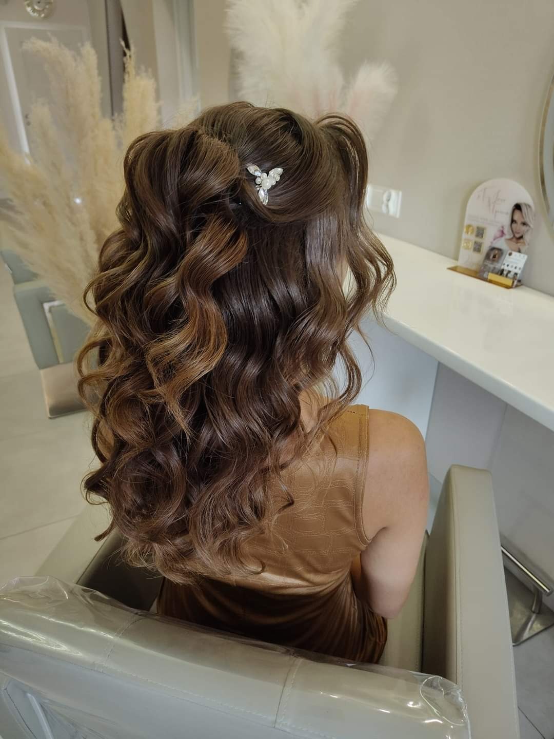 Upięte, falowane, brązowe włosy z refleksami, ozdobione srebrną spinką z perełkami, widok z tyłu w salonie.