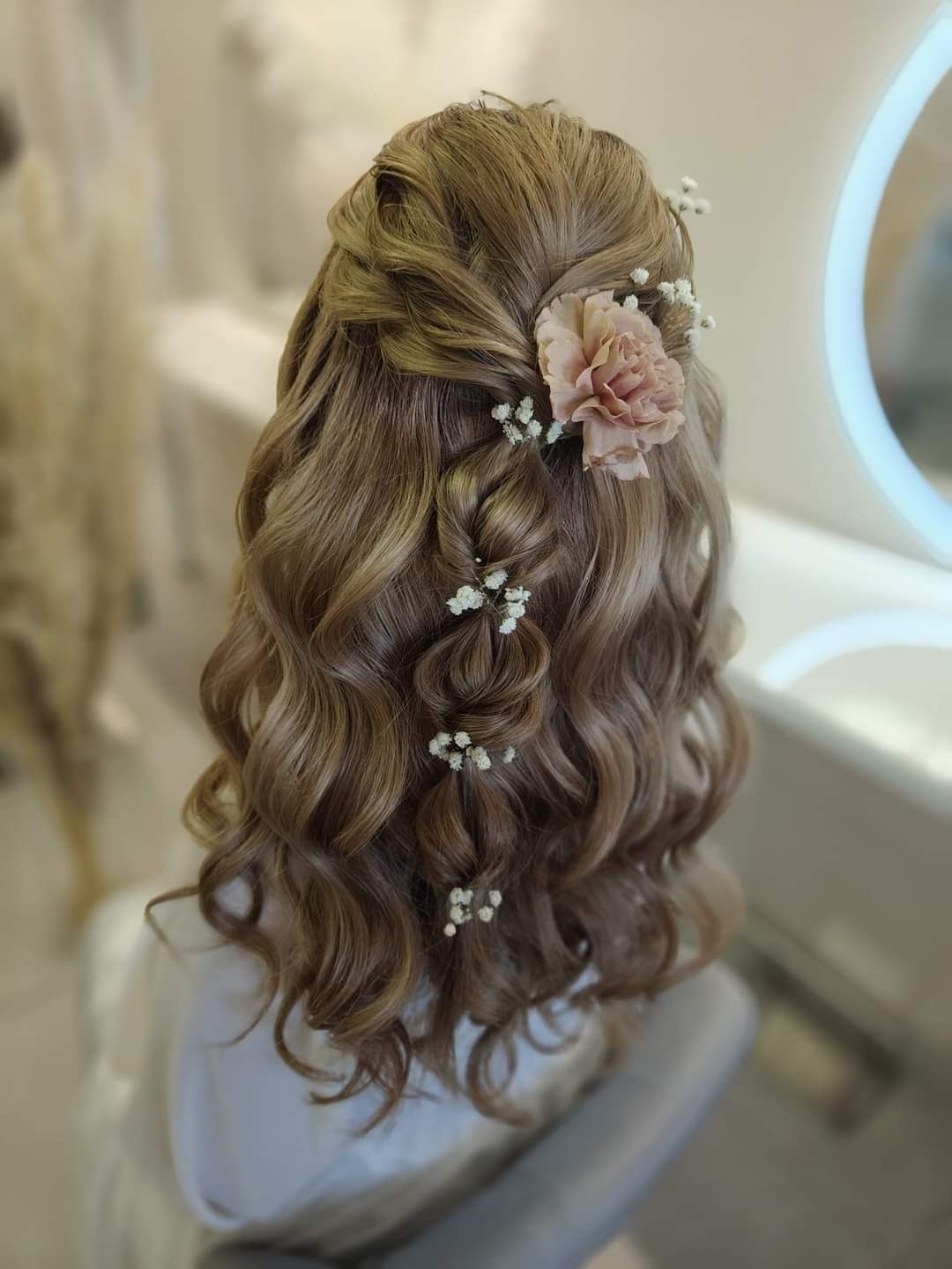Upięcie ślubne: delikatne fale w odcieniach blondu z wplecionymi warkoczami, ozdobione różowym kwiatem i drobnymi białymi kwiatuszkami.